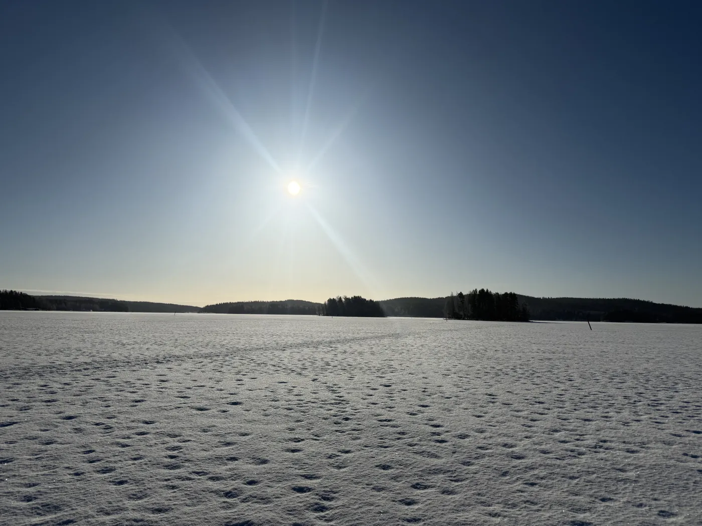 A frozen lake under a bright winter sun, snow-covered ice stretching to the treeline.